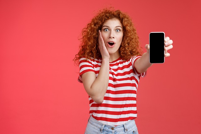 A young woman looks shocked as she holds up her smartphone, eyes wide and mouth open in surprise.